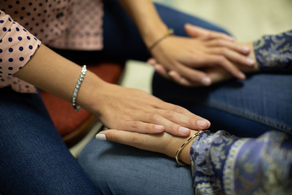 Simbolizando ThetaHealing: Uma mulher de blusa azul sentada com as mãos apoiadas nas coxas, e as palmas da mão viradas para cima. Enquanto outra mulher, sentada em uma cadeira na sua frente, está com as mãos sobre as palmas da mulher de azul.