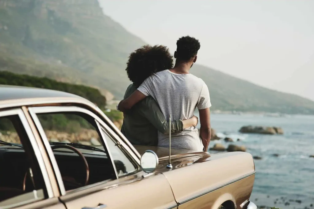 Casal sentado em capô de carro antigo, olhando para o mar, abraçados.