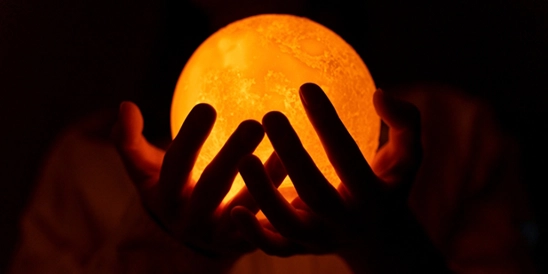 Mãos segurando luminária redonda, em formato de lua (luz amarela)