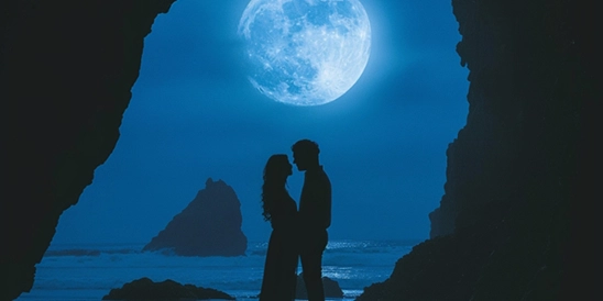 Casal, frente ao mar, sob a luz azulada da Lua Cheia. Só vemos silhuetas, o mar e o céu.