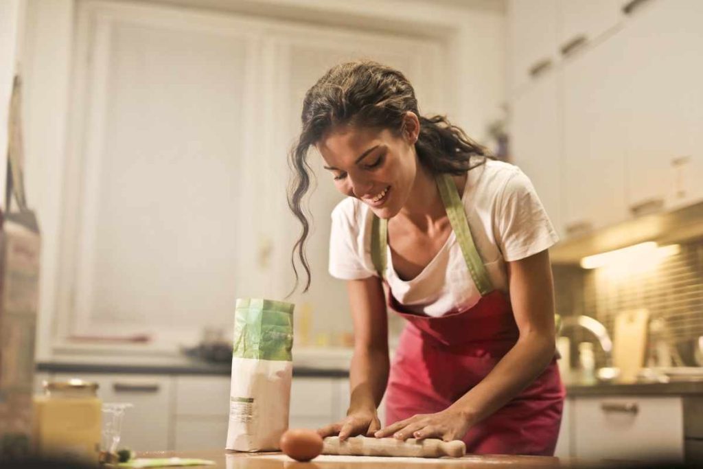 Mulher na cozinha, de avental, preparando uma massa e com um lindo sorriso no rosto.