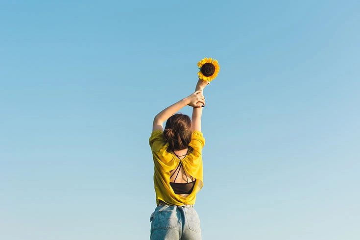 Mulher vestida de amarelo, de costas, segurando um girassol para o alto, em contraste com o céu azul