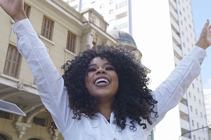 Mulher com camisa branca, braços levantados e sorriso no rosto, pela manhã em uma cidade.