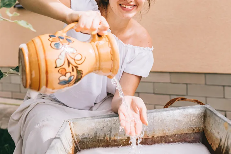 Mulher sorridente, despejando água de uma ânfora decorada em sua mão sobre uma tina de metal.