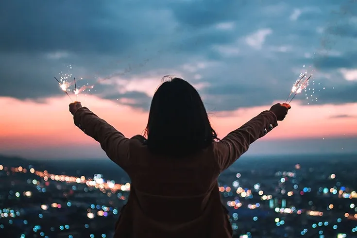 Mulher de braços abertos em um mirante. Ao longe, se vê uma cidade ao crepúsculo.