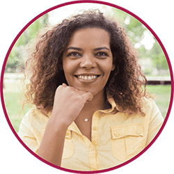 Imagem de uma mulher jovem sorridente com cabelo cacheado castanho, usando camiseta bege, com a mão apoiada no rosto, em fotografia de perfil circular.